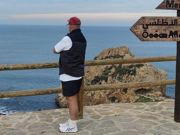 Man standing on a cliff-edge viewpoint overlooking the Atlantic Ocean with Arabic signpost