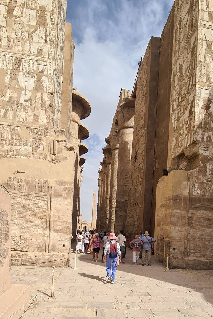 Row of monumental sandstone columns covered in hieroglyphs leading to distant obelisk under bright sky