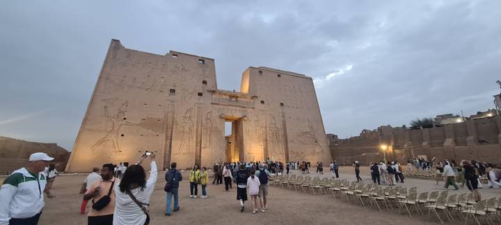 Evening tour group entering vast pylon gate of Edfu Temple lit by soft spotlights against cloudy sky