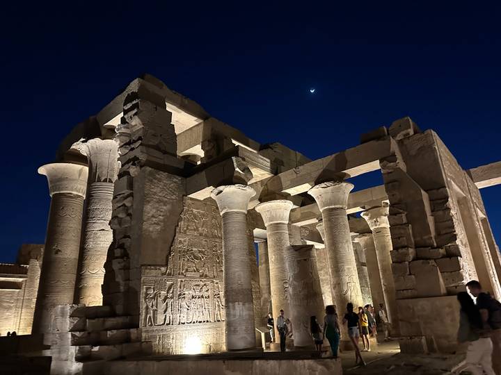 Beautifully lit columns and carvings of Kom Ombo Temple under a dark blue night sky with a crescent moon.