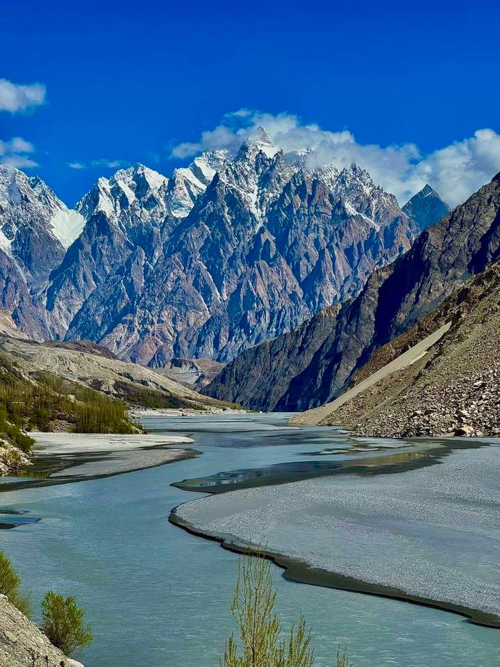 Dramatic snow-capped Karakoram peaks tower over a winding river in a steep rocky gorge.