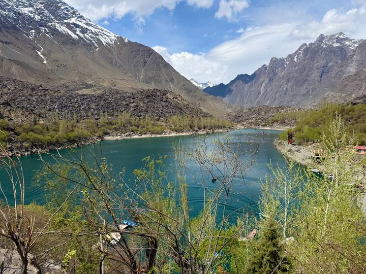 Turquoise alpine lake surrounded by budding trees and sheer rock faces of the Karakoram mountains.