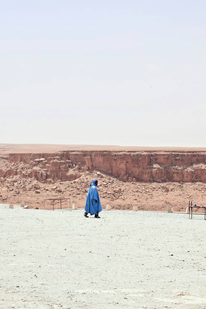A lone Berber man in a bright blue cloak stands before a rugged reddish cliff in harsh midday light.