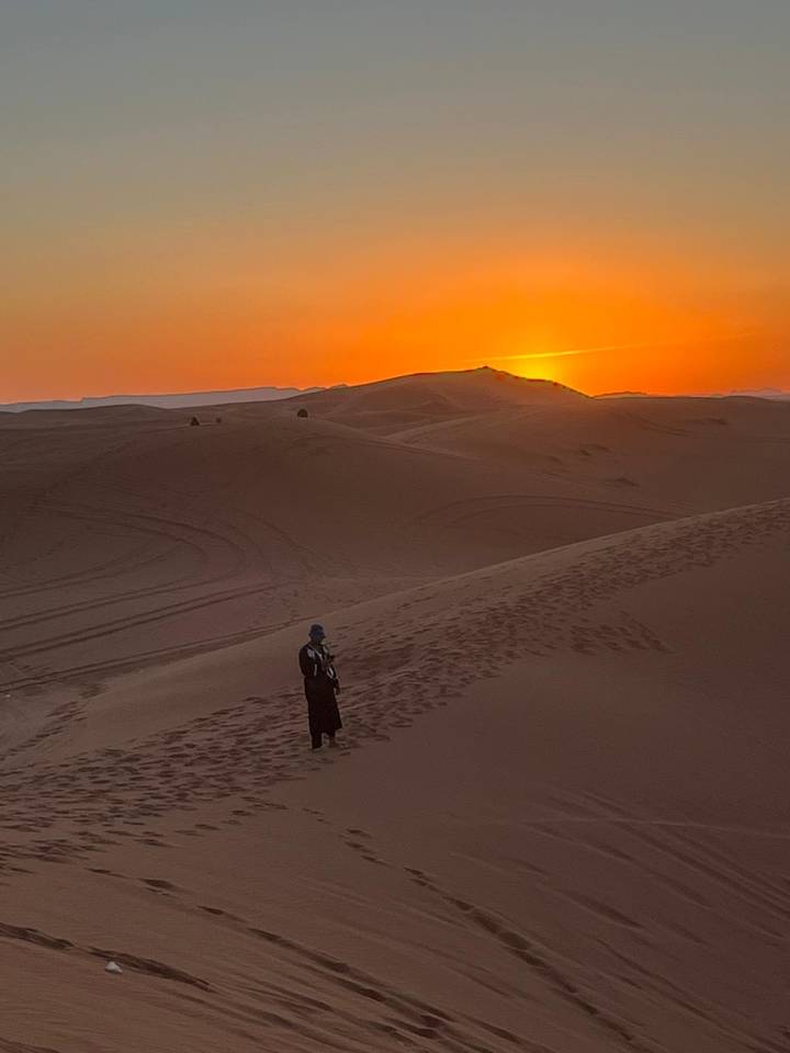 A lone traveler stands on a tall sand dune watching the orange sun set over rolling desert dunes with faint vehicle tracks.