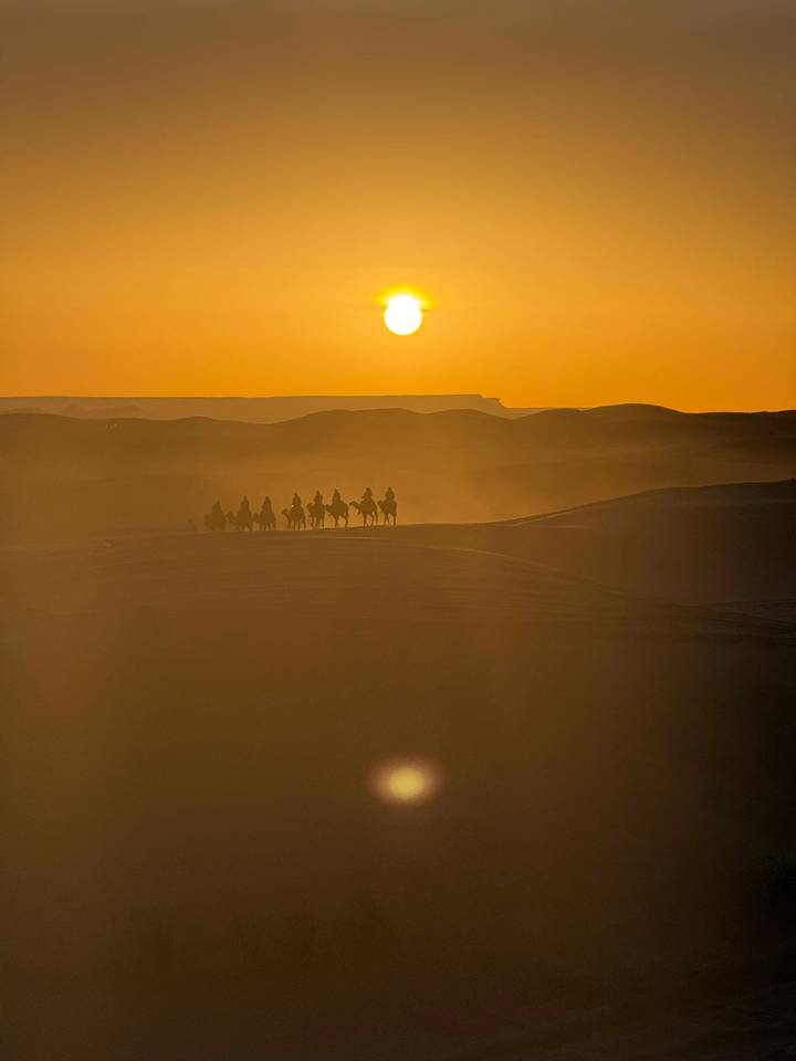A line of camel riders silhouetted against a glowing orange sunset, dust rising over sweeping desert dunes.
