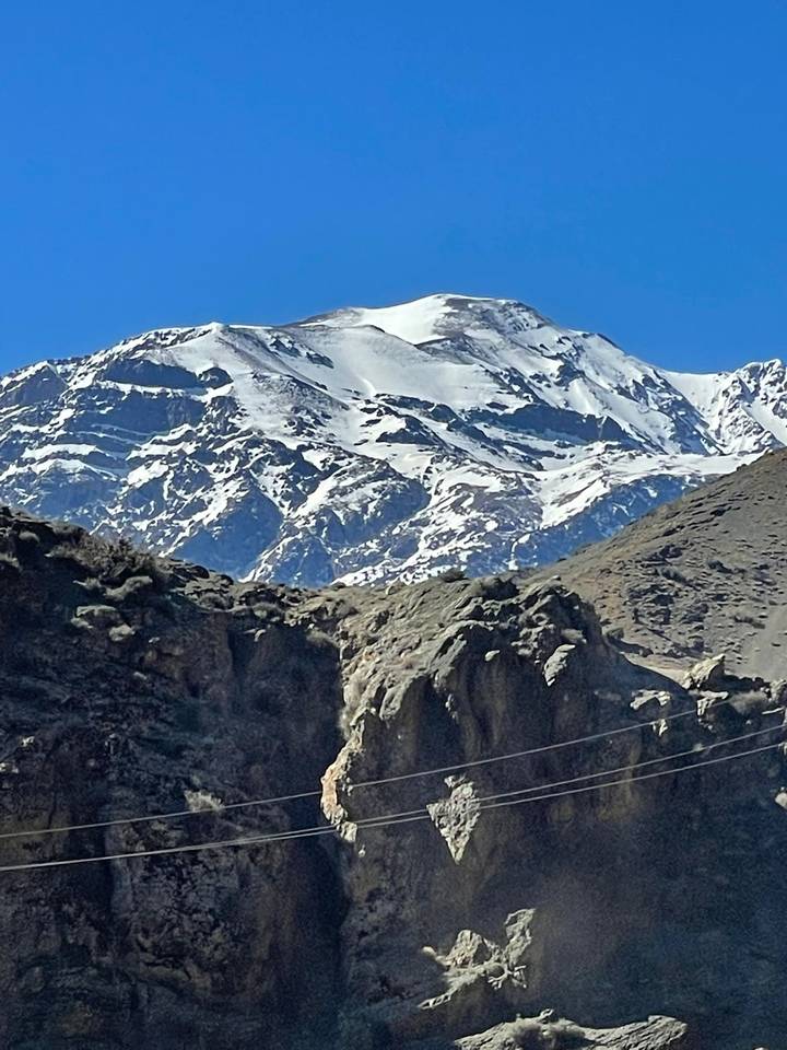 Snow-covered Atlas mountain peaks rise sharply above rugged rocky foothills under a clear sky.