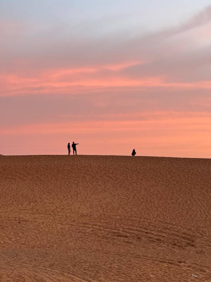 Three figures stand in silhouette on a dune against a vivid pink twilight sky.