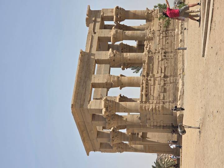 Ancient sandstone columns and capitals of an Egyptian temple rise from a sandy desert courtyard under a clear sky.
