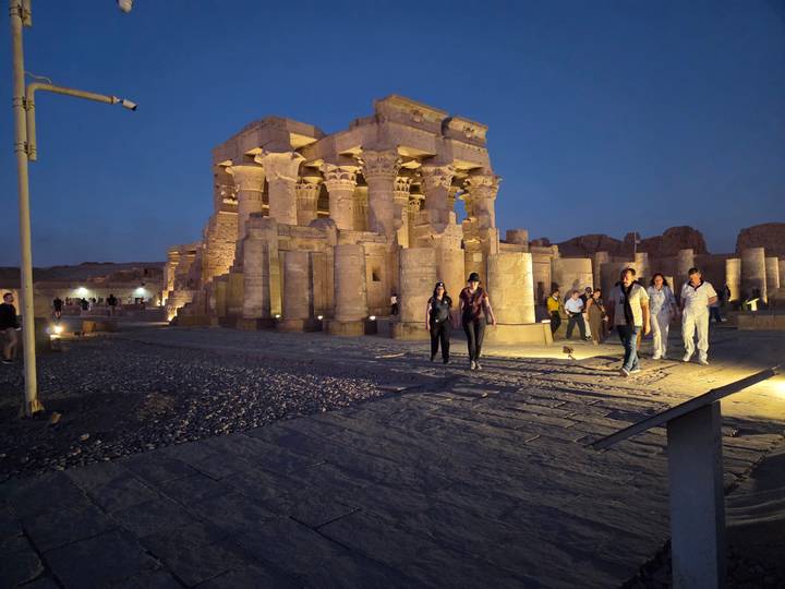 Nighttime view of a flood-lit Egyptian temple with tourists walking on the stone forecourt.