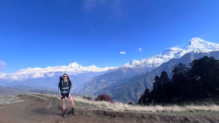 Female trekker with backpack stands on a ridge trail overlooking snowy Himalayan giants and blue sky.