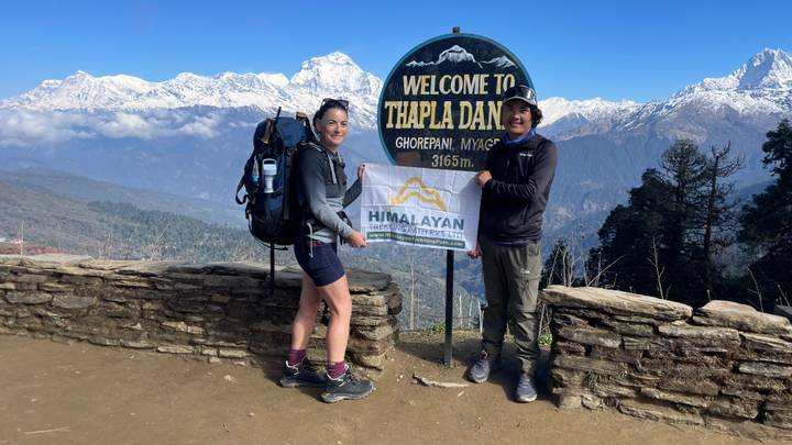 Trekkers smile beside a 'Welcome to Thapla Danda' sign with panoramic snowy peaks behind.