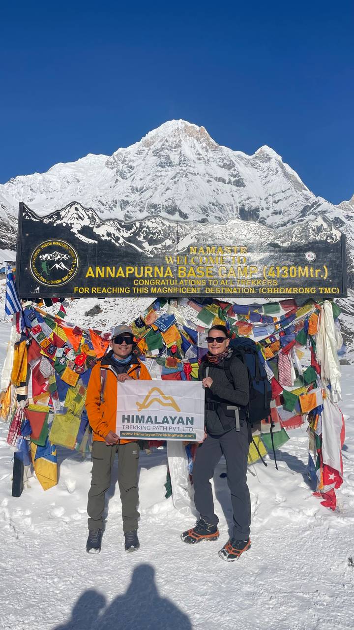 Close-up of two trekkers holding a banner in front of the Annapurna Base Camp welcome board covered in prayer flags.