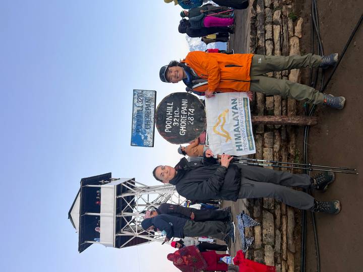 Early-morning photo of two trekkers with poles at the famous Poon Hill altitude sign and lookout tower.