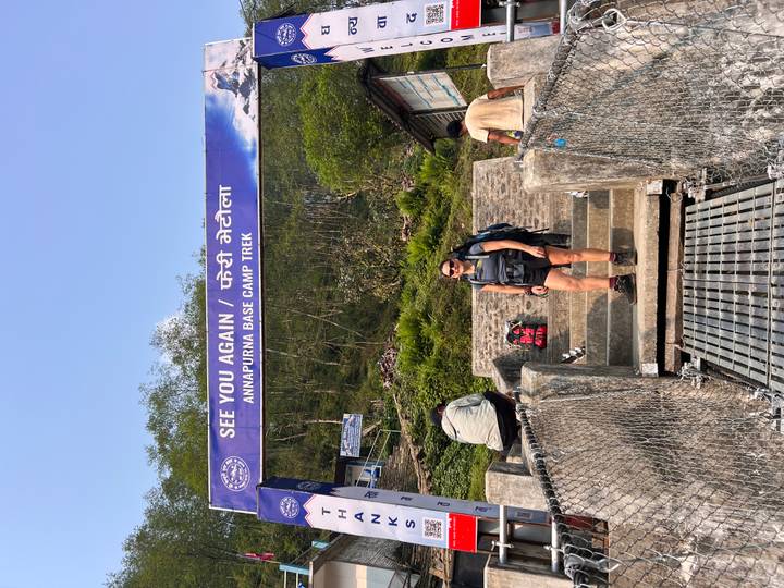 Trekker pauses under a farewell banner reading 'See You Again / Annapurna Base Camp Trek' at a suspension bridge exit.