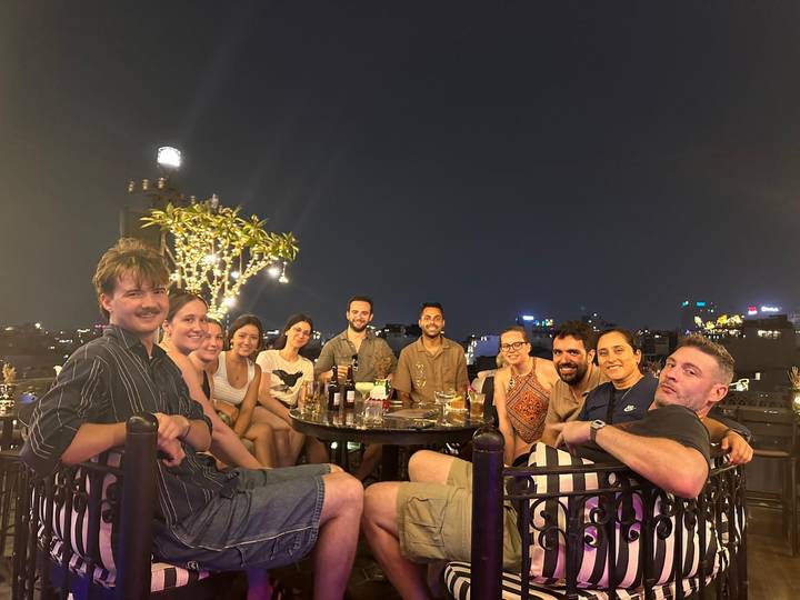 Smiling travellers share drinks around a rooftop table at night with a city skyline twinkling behind them.
