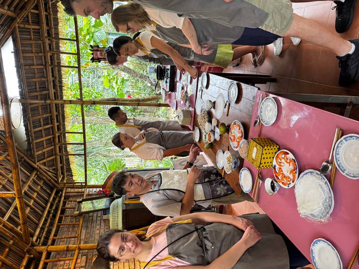 Travellers in aprons line a long table preparing ingredients during a hands-on Vietnamese cooking class in an open-air kitchen.