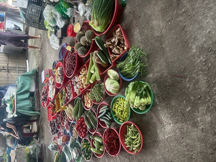 Array of fresh vegetables and spices displayed in red baskets on a market floor while shoppers browse.
