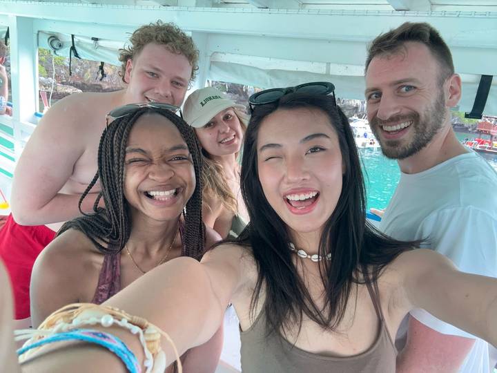 Group selfie on a boat with sparkling turquoise water and cliffs behind them in Thailand.