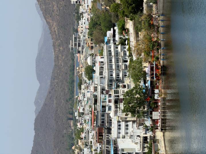 Panoramic view over white-washed buildings lining the lake in Udaipur with arid hills behind.