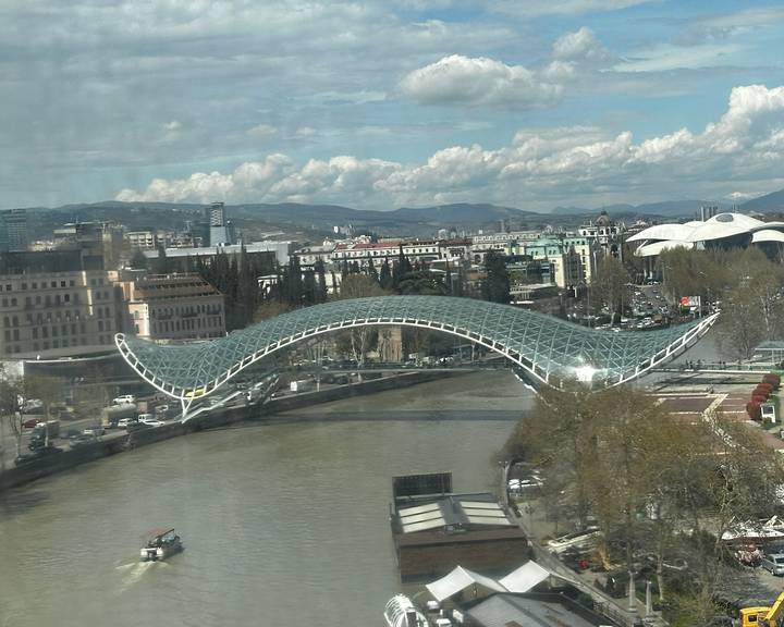 Modern arched glass pedestrian bridge spanning a river with Tbilisi cityscape and mountains in the background.