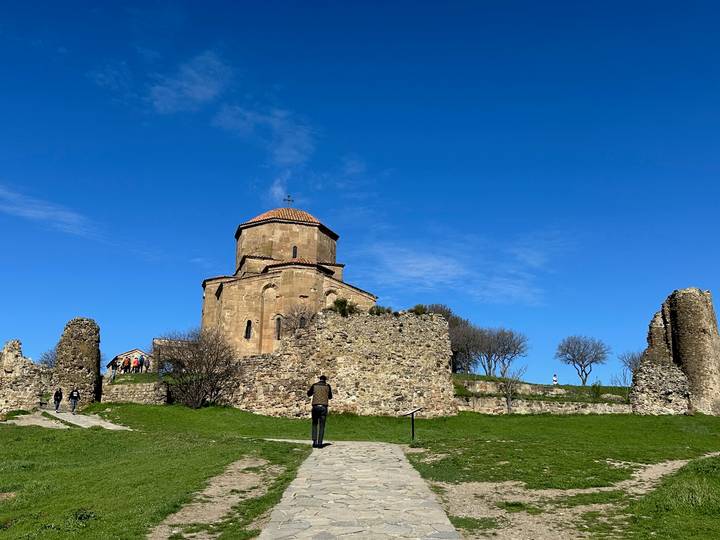 Ancient stone church and ruined fortress walls sit atop a grassy hill beneath a vivid blue sky, with a visitor walking the path.