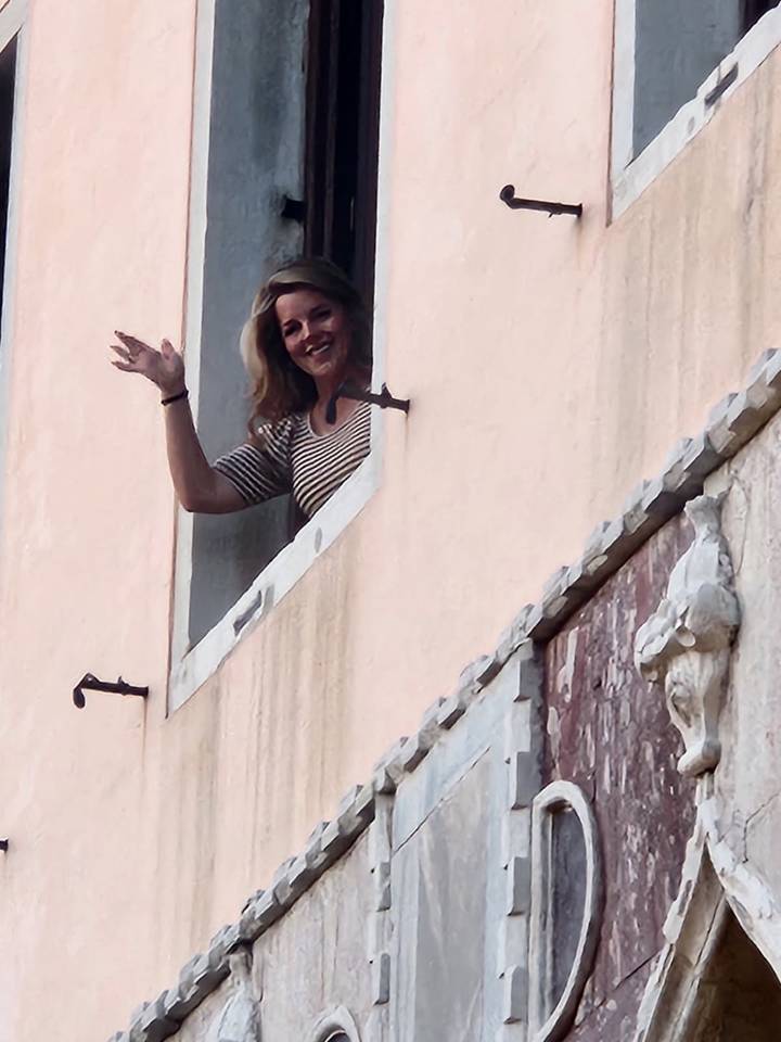 Smiling woman waving from an upper-storey window of a pastel stucco building