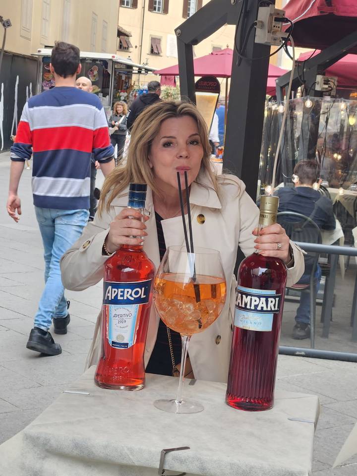 Woman humorously sipping a gigantic cocktail between oversized Aperol and Campari bottles on a busy street