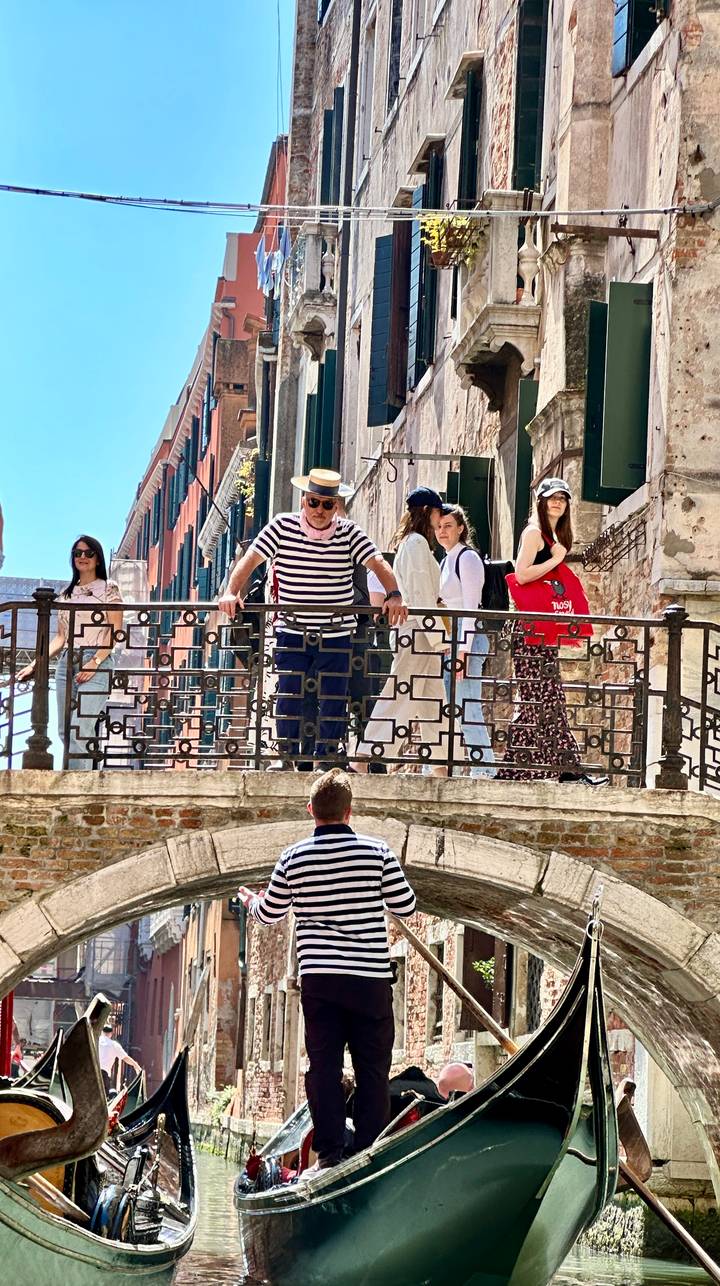 Striped-shirt gondolier leaning over an iron bridge while tourists watch along a Venetian canal