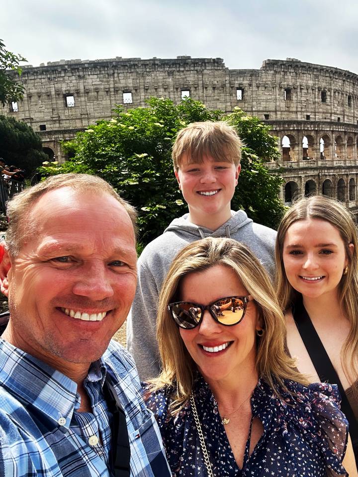Family selfie with the Colosseum partially visible behind lush greenery on a sunny day