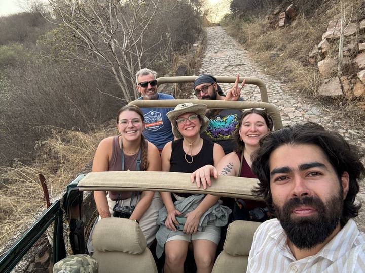 Friends take a cheerful selfie while riding in an open jeep along a rugged cobblestone trail lined with dry brush.