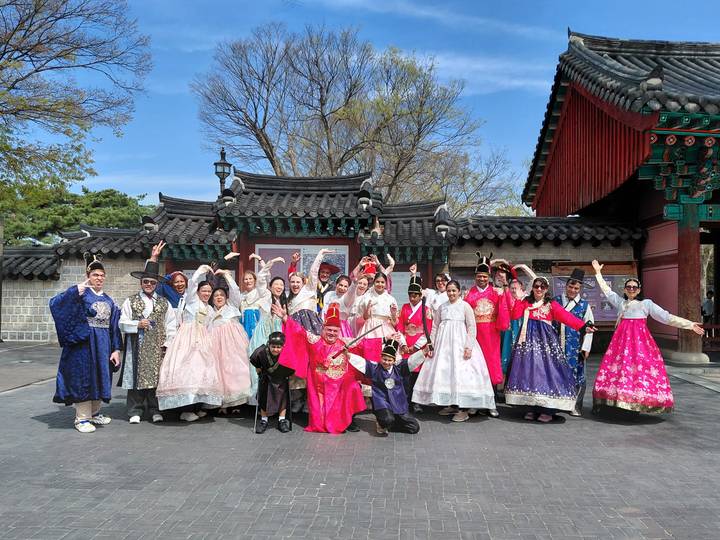 Large tour group dressed in vibrant traditional hanbok posing joyfully in front of an ornate palace gate.