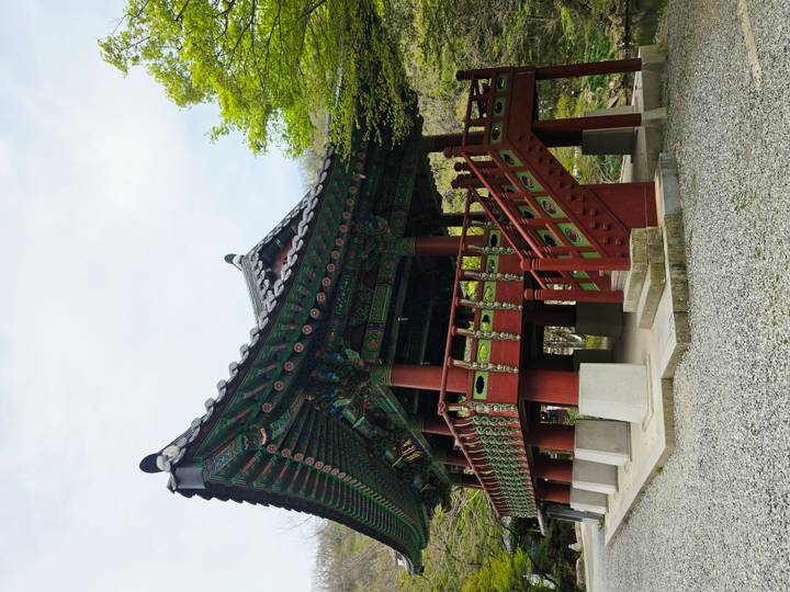 Colorfully painted wooden pavilion with ornate roof and red pillars set among greenery at a Korean temple site.