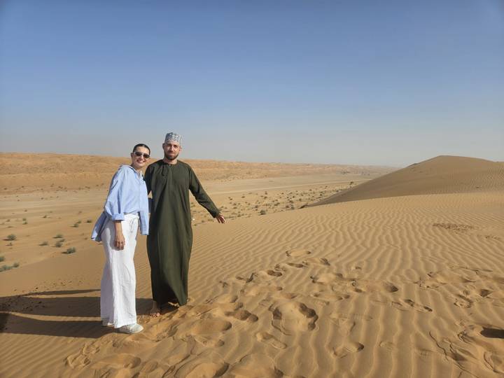 A couple poses atop rolling golden sand dunes under a clear sky in the desert.