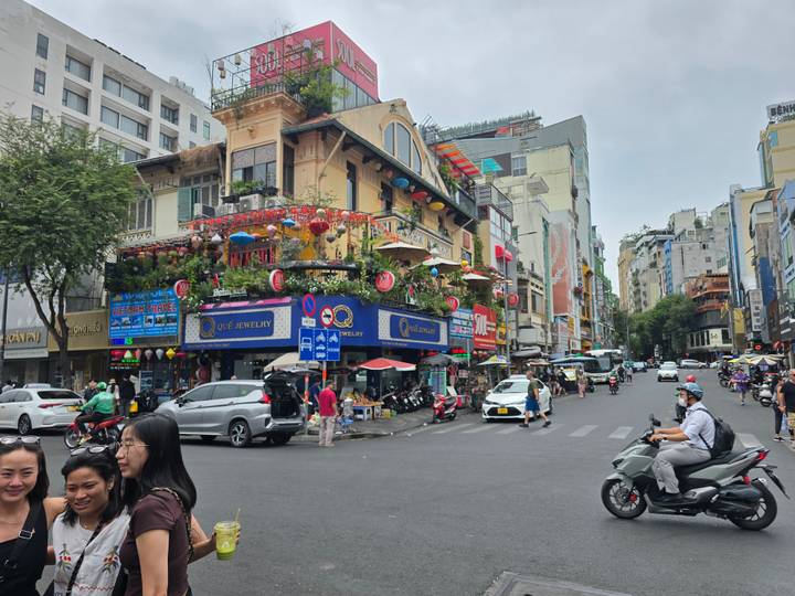 A lively city intersection bustles with scooters, pedestrians, and colorful shopfronts decorated with plants and lanterns.