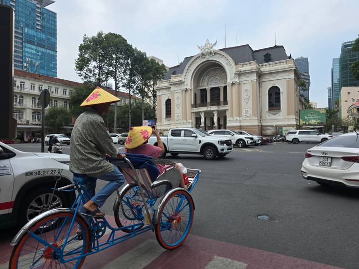 A traditional cyclo with passengers wearing conical hats passes the ornate Saigon Opera House.