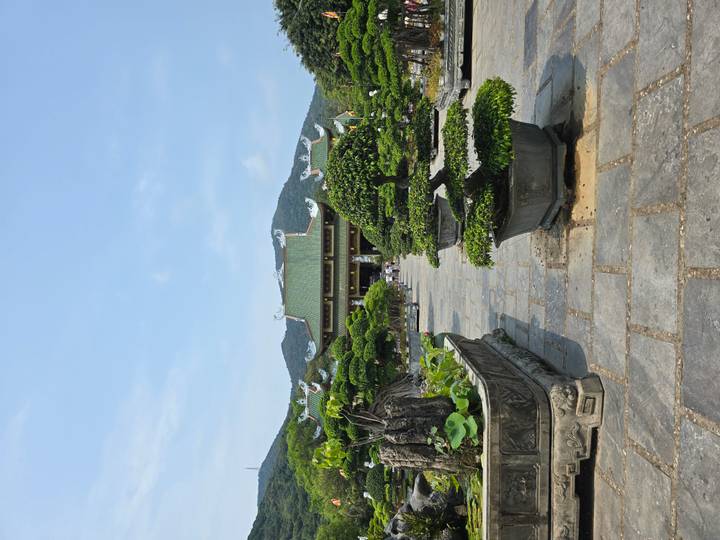 A bonsai-filled courtyard leads to a green-tiled temple set against forested mountains.