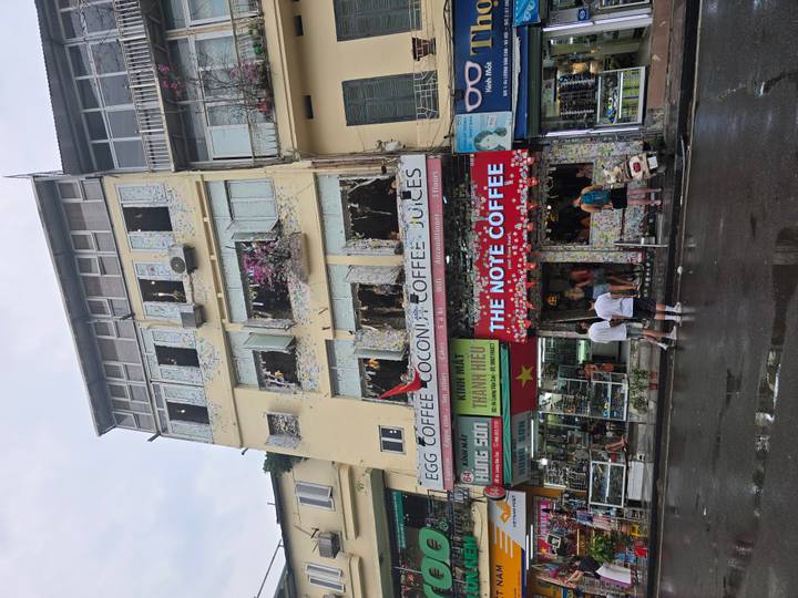 A colorful coffee shop plastered with sticky notes stands on a busy Hanoi street after rain.