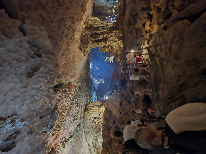Tourists explore a vast illuminated cave chamber with stalactite formations and rocky paths.