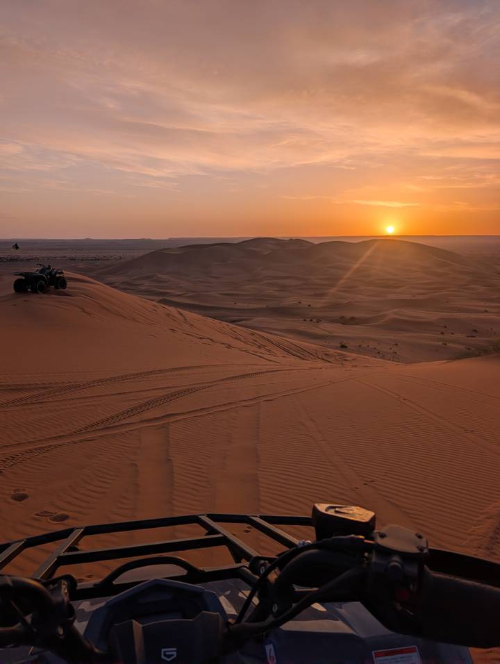Sun setting over rolling sand dunes with quad-bike tracks and a lone vehicle silhouetted on a ridge.