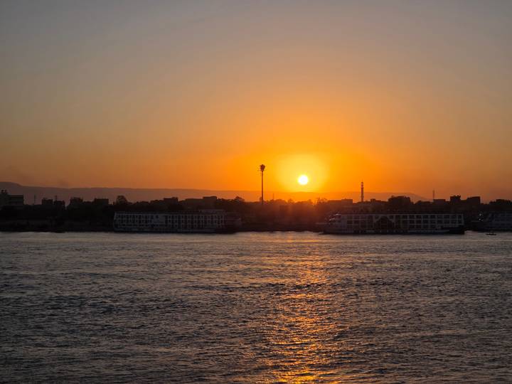 Orange sun setting behind silhouette of riverside cityscape along wide calm river.