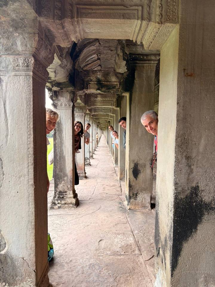 Smiling travellers peek out between stone columns in a long Angkor temple corridor.