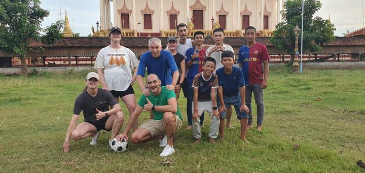 Travellers and local youths pose with football on grassy field beside ornate stupa.
