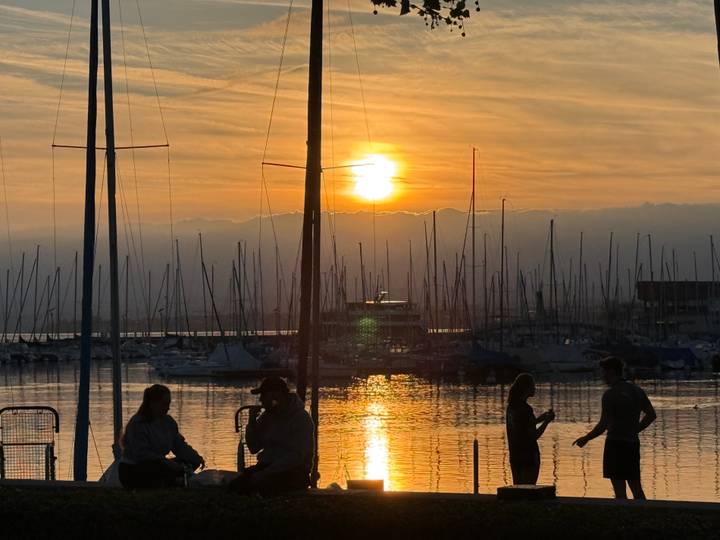 Silhouetted figures chat on a pier as the sun sets over a marina filled with sailboat masts.