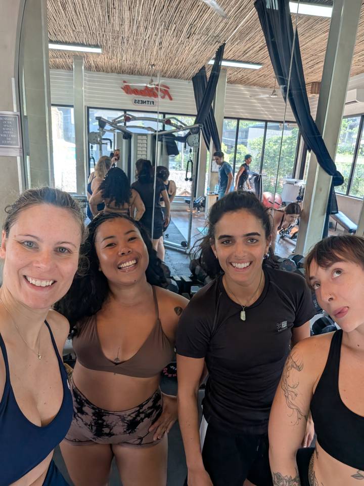 Gym selfie with four women smiling after a workout, exercise equipment and mirrors behind.