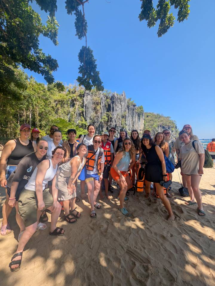 Smiling tour group posing on a sandy beach with dramatic limestone cliffs behind.