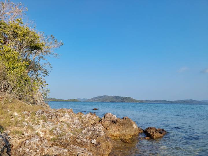 Rocky shoreline with clear blue water and distant rolling hills under bright sky.