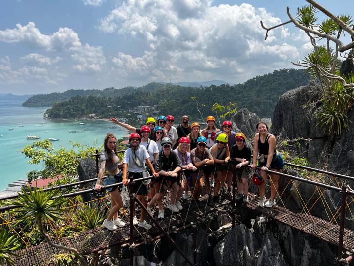 Energetic helmeted group posing on a cliff bridge with turquoise bay backdrop.