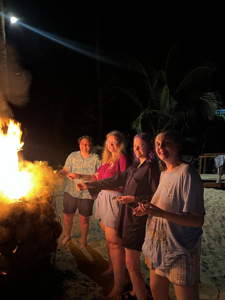 Four friends roast marshmallows over a bright beach bonfire at night, smiling at the camera with palm trees behind them.