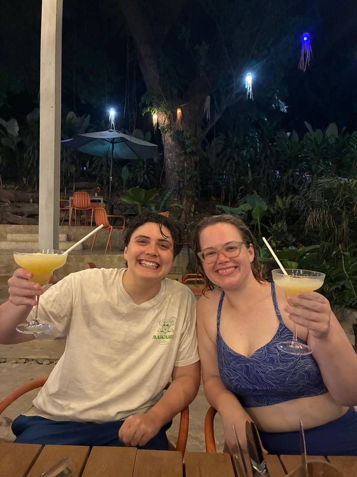 Two travellers toast frozen cocktails at an open-air bar surrounded by tropical plants at night.