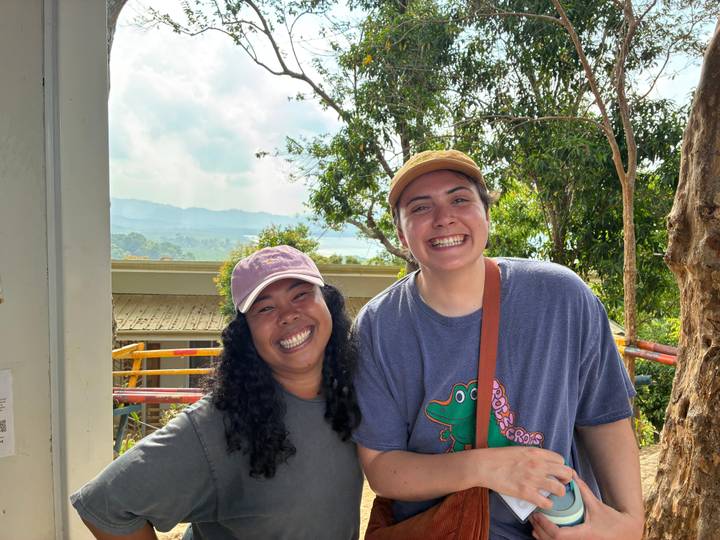 Two smiling women pose outdoors with rolling hills and misty mountains in the background on a sunny day.